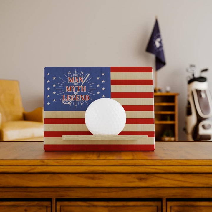 Golf ball on a stand with American flag design and 'Man Myth Legend' text in a room setting.