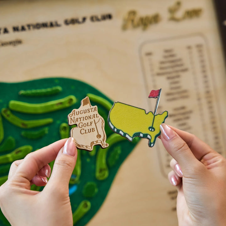 Two wooden pins of the United States and Augusta National Golf Club held by a person against a blurred background.