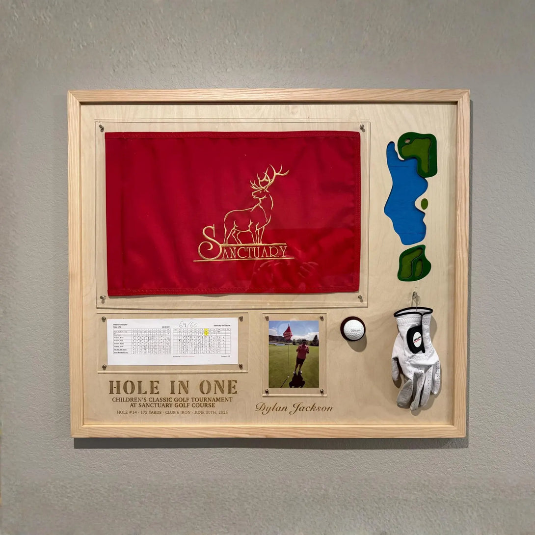 Framed golf-themed display with red flag, golf glove, and ball on a gray wall.